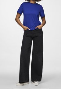 Persona che indossa una camicia blu a maniche corte, pantaloni ampi neri a vita alta e sneakers platform bianco e nere, con le mani in tasca.