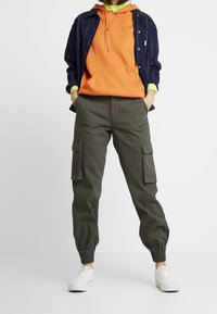 Henkilö, jolla on yllään oliivinvihreät cargo-housut, joissa on joustavat hihnat, valkoiset tennarit, oranssi huppari ja tummansininen raitaovershirt.