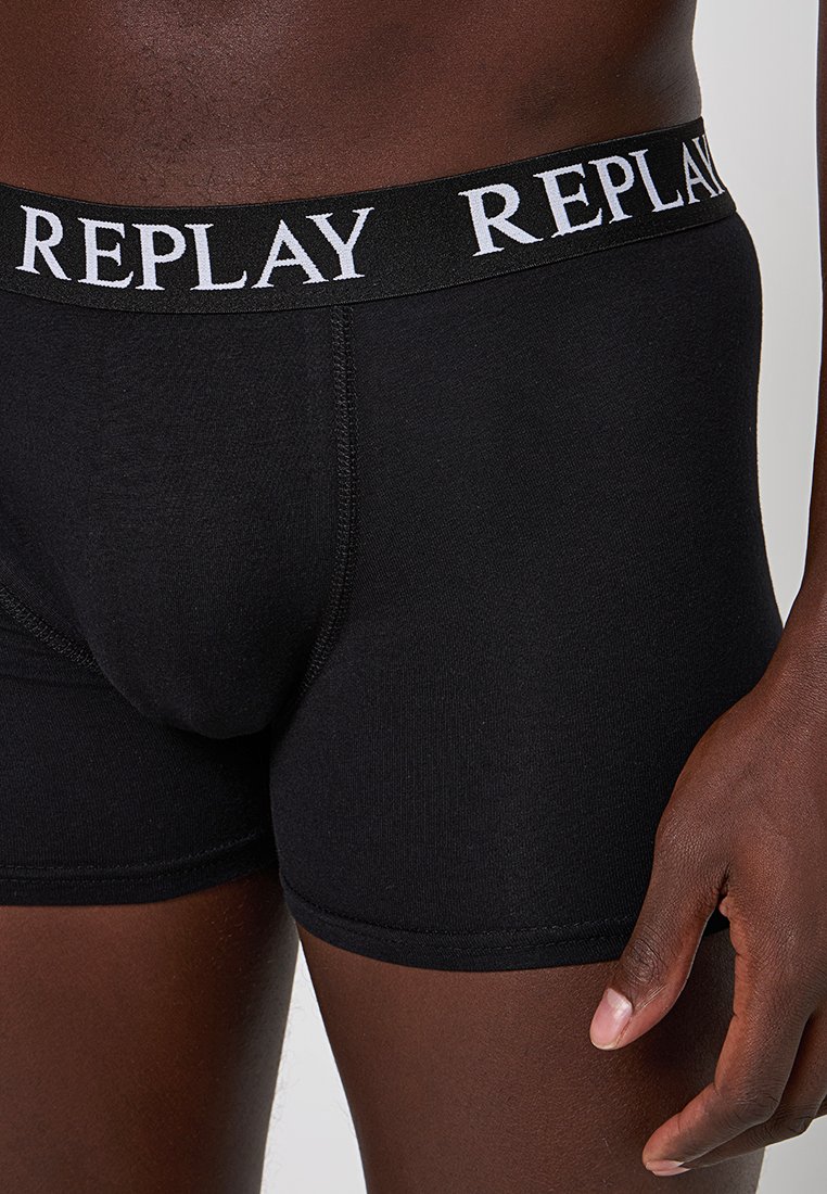 Nahaufnahme einer Person, die schwarze Boxershorts mit einem schwarzen Bündchen trägt, das mit weißem "REPLAY"-Branding versehen ist.