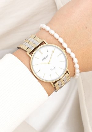 Montre ronde avec un cadran blanc, boîtier en métal doré et bracelet en maillons mélangés argent et or. Accompagnée d'un bracelet en perles blanches.