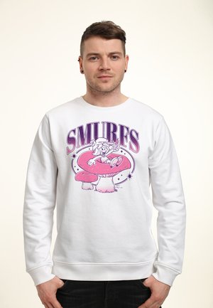 Mand iført hvid sweatshirt med lilla tekst "SMURES" og tegneseriefigur, der sidder på en stor lyserød svamp.