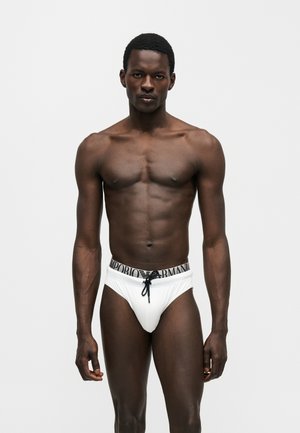 Μυώδης σκουρόχρωμος άνδρας όρθιος, φορώντας λευκό μαγιό Emporio Armani με μαύρο κορδόνι, σε απλό γκρι φόντο.