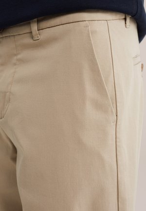 Lähikuva beigejen housujen taskusta ja vyölenkistä, jotka on yhdistetty tummaan yläosaan, näyttää kankaan tekstuurin ja sauman yksityiskohdat sivulla.