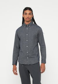 Tânăr cu împletituri lungi, purtând o cămașă gri cu butoni și pantaloni negri, stând pe un fundal alb simplu.