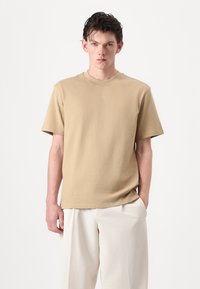 Beige Baumwoll-T-Shirt mit kurzen Ärmeln und rundem Ausschnitt, mit einem dezenten "HUGO"-Logo auf der Brust. Glatte Textur, lockere Passform.
