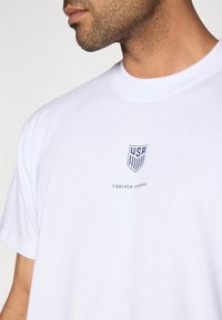 T-shirt de algodão branco com um logótipo azul do escudo dos EUA e o texto "FOREVER FORWARD" impresso por baixo numa fonte simples sem serifa.