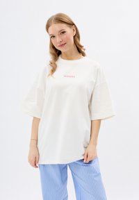 T-shirt en coton blanc à manches courtes et coupe décontractée, avec un petit logo en texte rose "MIRONS" sur le devant. Pantalons rayés bleus.