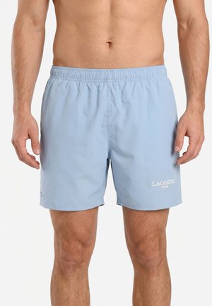 Lichtblauwe Lacoste zwemshorts met elastische taille, gedragen door een man, met logo op het linkerbeen.