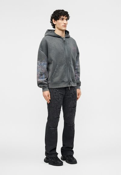 Muž s kučeravými vlasmi, oblečený v sivom pulóvri so zipsom s textom na rukávoch, čiernych potrhaných džínsoch a čiernych teniskách, stojaci čelom vpred.