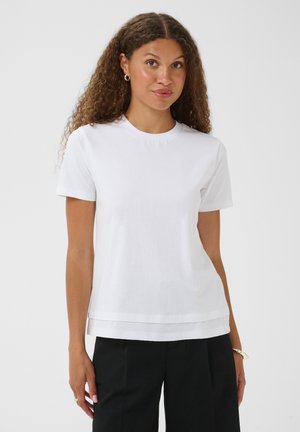 Femme aux cheveux bouclés portant des boucles d'oreilles argentées en forme d'anneau, un t-shirt blanc uni à manches courtes et un pantalon noir sur un fond uni.