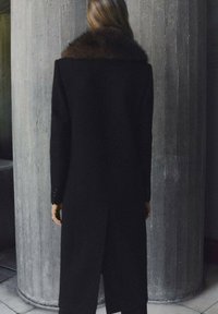 Abrigo largo negro con cuello de piel marrón, diseño estructurado, textura suave y ajuste a medida, mostrando un estilo minimalista contra un fondo de hormigón.