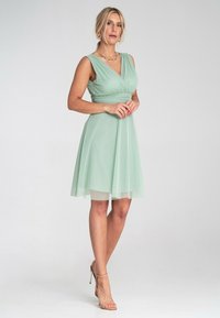 Mintgrünes, ärmelloses Kleid mit V-Ausschnitt und gerafftem Bund. Der Rock ist luftig und endet oberhalb des Knies, mit einem transparenten Überzug. Zarte Textur.