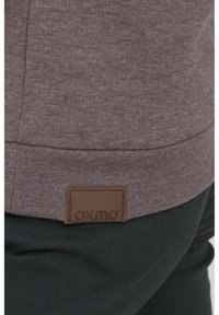 Brun sweatshirt med texturerad yta, med en läderetikett som har "oxmo" vid nederkanten, i kombination med svarta byxor.