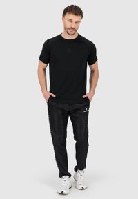 Schwarzes sportliches T-Shirt mit kurzen Ärmeln und rundem Halsausschnitt, kombiniert mit schwarzen Hosen, die seitliche Logos haben, abgerundet mit weißen Sneakers.