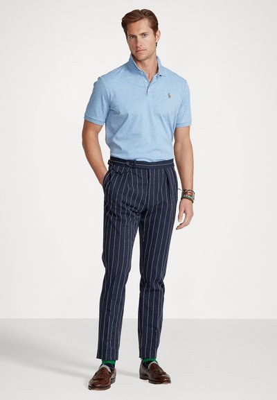 Világoskék pólóing gallérral, tengerészkék csíkos nadrág és barna mokaszinok. Figyelemre méltó zöld zoknik láthatóak, karcsú szabás.