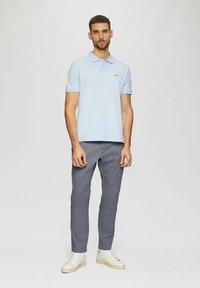 Hellesblaues Polohemd mit kurzen Ärmeln, graue Hose und weiße Sneaker. Das Polohemd hat ein dezentes Logo auf der Brust.