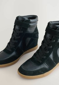 Sneakers alte in suede verde scuro e pelle nera. Le caratteristiche includono chiusura con lacci, cinturino alla caviglia, punta perforata e suola in gomma.