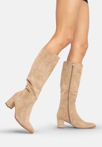 Botas de ante beige hasta la rodilla con tacón cuadrado, que cuentan con un zipper lateral y una textura suave, diseñadas para ajustarse cómodamente alrededor de la pantorrilla.