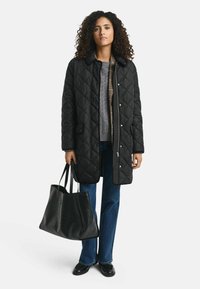 Zwarte gewatteerde lange jas met drukknoopsluiting, voorzien van een hoge kraag. Gecombineerd met een grijze trui, blauwe flared jeans en een zwarte gestructureerde tote bag.