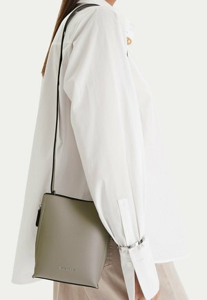 Osoba v bílém tričku s dlouhým rukávem a béžových kalhotách, nesoucí šedou kabelku přes rameno s černým popruhem.