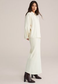 Vrouw met lang donker haar, gekleed in een ivoorkleurige blouse met lange mouwen, een bijpassende rok tot de enkels en donkerbruine hakken, staande in profiel.