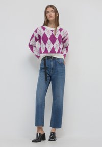 Femme portant un pull en argyle violet et blanc, un jean taille haute bleu avec une ceinture noire, et des chaussures à talons noires, se tenant contre un mur blanc.