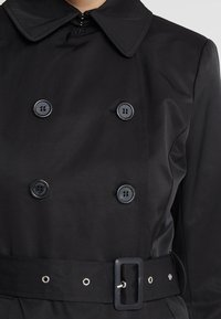 Černý dvouřadý trenčkot se čtyřmi knoflíky a širokým pásem s obdélníkovou sponou, zobrazený od hrudníku po pás.