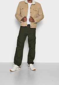 Man in een beige jas met sherpa-kraag, een wit T-shirt, olijfgroene cargopants en witte sneakers staat tegen een effen achtergrond.