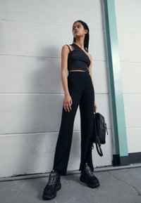 Débardeur noir asymétrique court, pantalon noir taille haute à jambes larges, et baskets noires épaisses. Sac à dos noir porté sur une épaule.