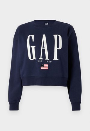 Tengerészkék rövidített pulóver fehér "GAP" felirattal, "EST. 1969" szöveggel és egy kisméretű, 13 csillagos amerikai zászlóval a szöveg alatt.