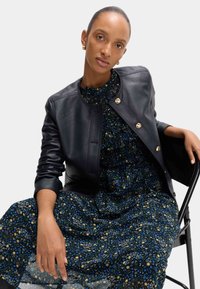 Czarna skórzana kurtka bombowca z złotymi guzikami na sukience w kwiatowy wzór z niebieskimi, żółtymi i białymi kwiatami na ciemnym tle.