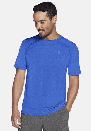 Mann trägt blaues kurzärmeliges Sportshirt und graue Shorts, schaut nach unten, eine Hand in der Hosentasche.