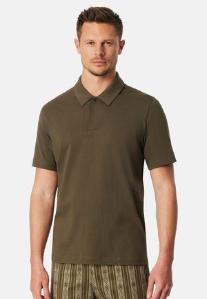 Muž v olivově zelené polokošili s krátkým rukávem a vzorovaných hnědo-zelených kalhotách stojí na bílém pozadí.