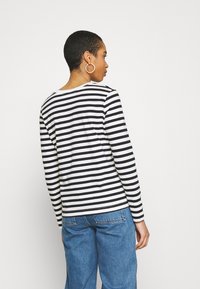 Tricou cu dungi alb-negru, cu mânecă lungă și decolteu rotund, asortat cu blugi albaștri din denim, văzut din spate.