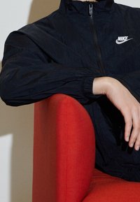 Nike vindjacka i svart, lättviktigt tyg med dragkedja fram. Har hög krage och elastiska ärmmuddar. Poserad mot en röd stol.