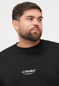 Schwarzes Baumwoll-T-Shirt mit Rundhalsausschnitt, das auf der Brust das weiße Textlogo "JJ Studios" und eine glatte, einfarbige Textur aufweist.