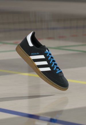 Čierne tenisky Adidas Spezial s bielymi pruhmi, modrými šnúrkami a gumovou podrážkou, ktoré sa vznášajú nad podlahou telocvične s farebnými čiarami ihriska.