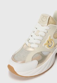 Beige og hvite sneakers med metalliske detaljer, mesh-paneler og en strukturert såle. Har hvite lissser og en logo på tungen.