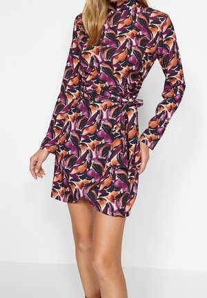 Kobieta ubrana w mini sukienkę z długim rękawem i wysokim dekoltem, ozdobioną abstrakcyjnym, fioletowo-pomarańczowo-czarnym kwiatowym wzorem oraz przewiązaną w talii.