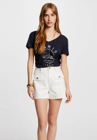 Camiseta de cuello en V azul marino con estampado de "París", combinada con pantalones cortos blancos de talle alto que cuentan con detalles de botones y grandes bolsillos frontales.