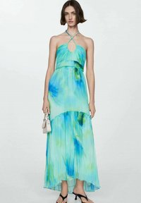 Maxi haljina, plisirani materijal u aqua i zelenom tie-dye uzorku, halterneck izrez s dekolteom, lepršavi siluet, u kombinaciji s crnim sandalama.