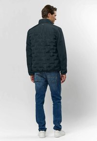 Homme portant une veste matelassée noire, un jean bleu et des baskets blanches, debout dos à un fond clair uni.