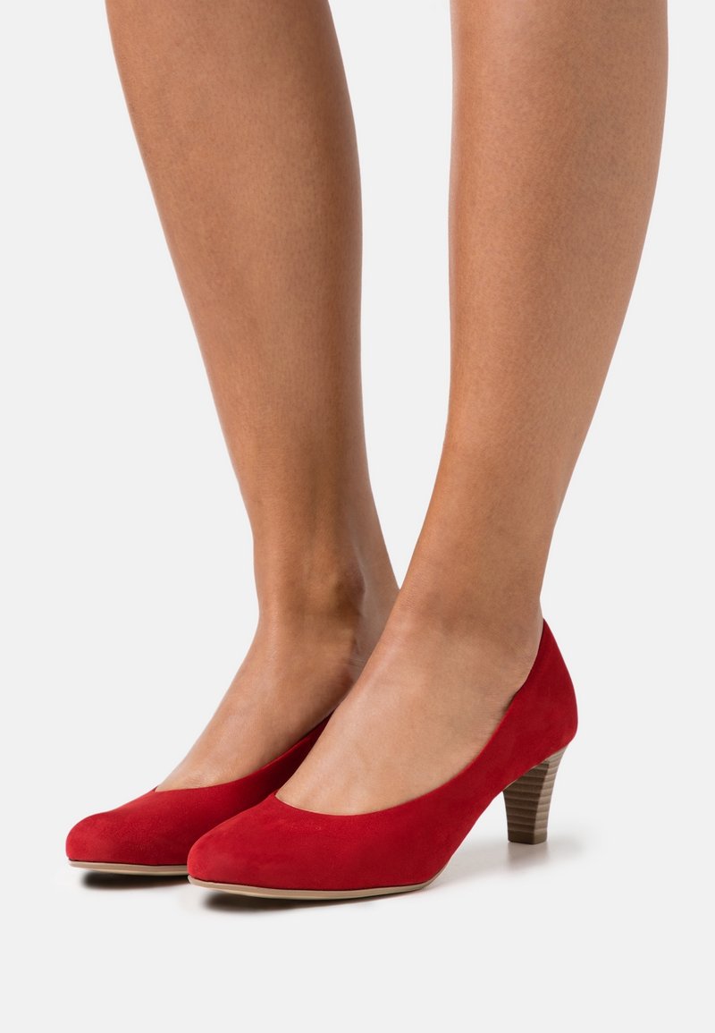 Jambes de femme portant des escarpins en daim rouge à talons moyens et bouts arrondis sur un fond clair uni.