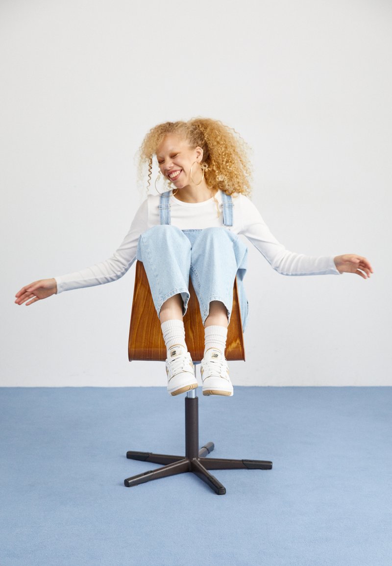 Chaise en bois avec une base ronde, présentant une personne en salopette bleu clair et chemise blanche, portant des baskets blanches, assise confortablement.