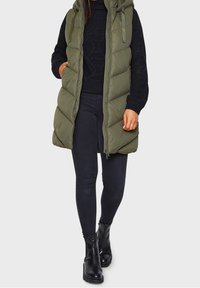 Olive green puffer vest with a hood, zip closure, and quilted design, worn over a black sweater and paired with black skinny jeans and ankle boots.