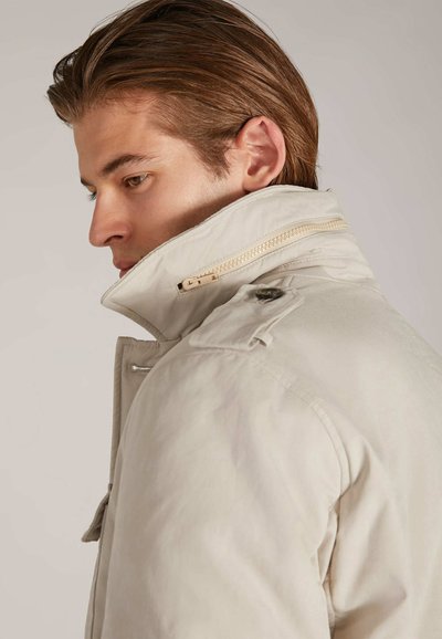 Beige jas met hoge kraag, voorzien van een ritssluiting en knoopaccenten. Lichtgewicht stof met een gladde textuur en een getailleerde pasvorm.