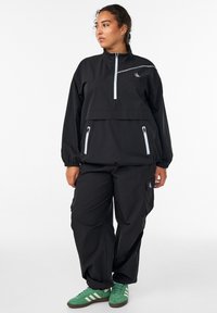 Black windbreaker with half-zip, elastic cuffs, and two zippered pockets. Paired with baggy black pants and green sneakers with white stripes.