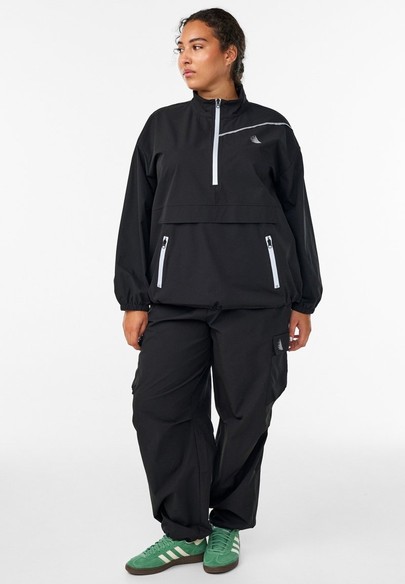 Black windbreaker with half-zip, elastic cuffs, and two zippered pockets. Paired with baggy black pants and green sneakers with white stripes.