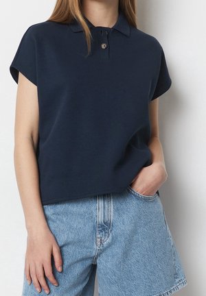 Femme portant un polo bleu marine à manches courtes et un short en denim bleu clair taille haute, une main dans la poche, devant un mur blanc.