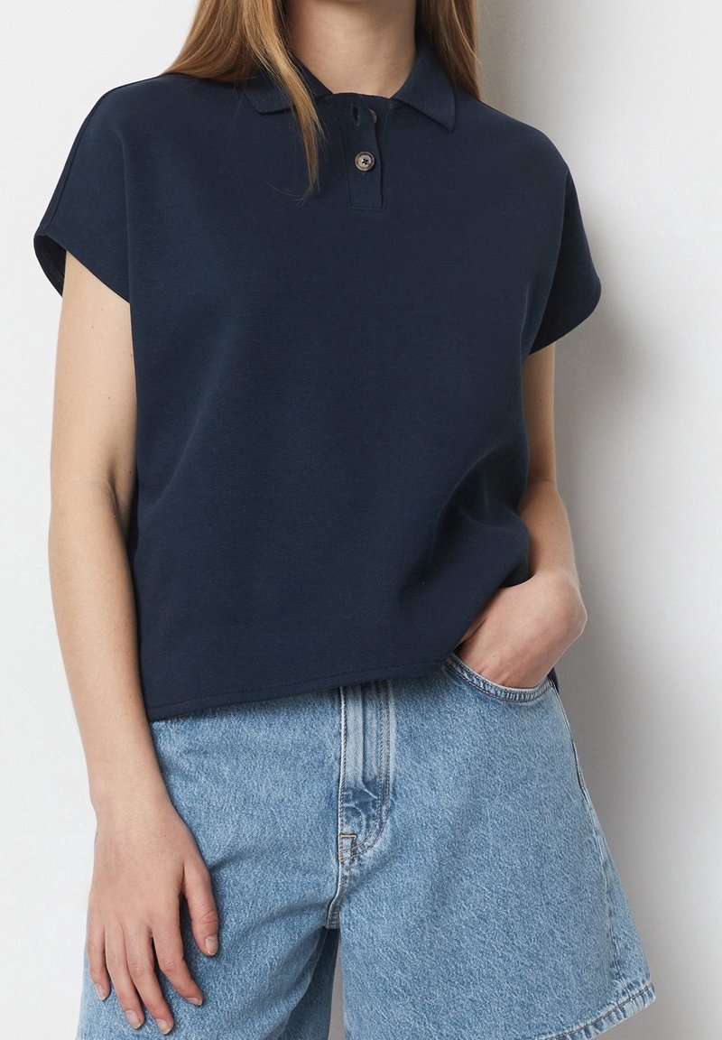 Femme portant un polo bleu marine à manches courtes et un short en denim bleu clair taille haute, une main dans la poche, devant un mur blanc.